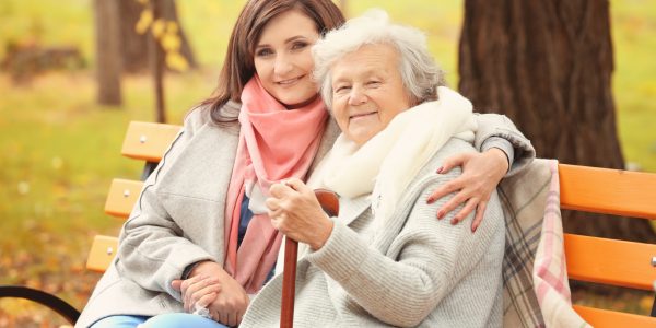 Senior woman with cane and young caregiver sitting on bench in park