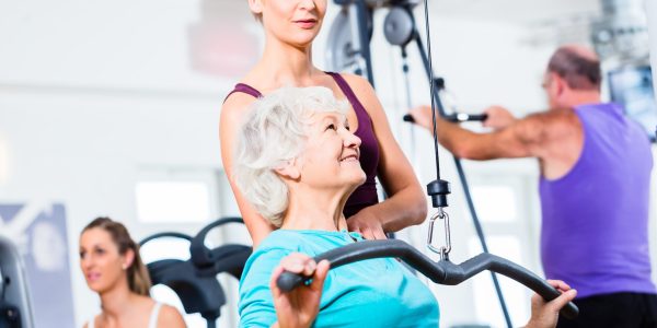 Senior woman doing back training with trainer in gym at machine